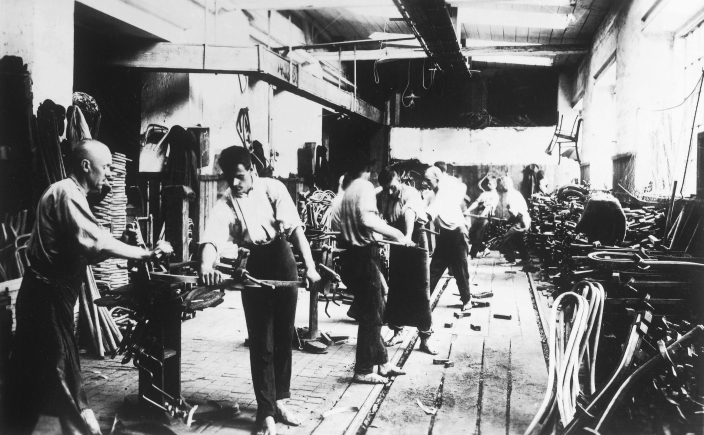 Thonet工場・曲木作業（約1920年）。曲木椅子の部材を人が治具で曲げる工場の記録写真。約1920年とされ、1859年のNo.14設計時の現場写真ではない。写真から蒸気の温度、型の寸法、個々の作業者の労働条件は読めない。