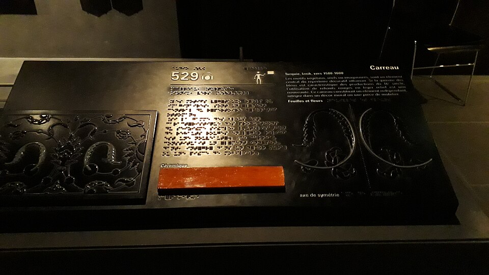 ルーヴルの触知・点字案内。2019年撮影。イスラーム美術部門の展示解説盤には、凹凸のある装飾図、点字、大きな文字が同居する。写真は装置の形を示すが、実際の読み方や利用者の評価は示さない。